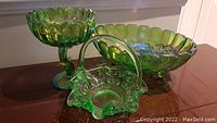 Three vintage green glass pieces, including a large oval bowl with harvest pattern, a pedestal bowl, and a basket with handle arranged on wooden surface.