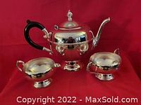 Electroplated silver coffee pot, cream jug and sugar bowl with black handle on coffee pot and ornate detail on spout and handles photographed against red backdrop.