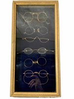 Full view of the sealed framed display showing all six pairs of antique eyeglasses vertically arranged on dark backing inside a gold decorative frame.