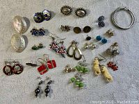 Full top-down view showing all earrings laid out on fabric. Shows variety and condition of earrings with some having shiny enamel and others pearl accents.