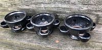 Full view of the set of three ceramic strawberry planter pots placed on a wooden surface, showing their glossy dark gray gunmetal finish, rounded shape, and handles.