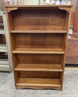 Front view of the wooden bookcase showing four shelves and medium brown finish. Decorative brackets visible on the top section.