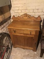 Front view of antique wooden washstand against a brick wall, showing drawer with handles and double-door cabinet below.
