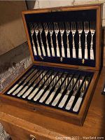 Full view of opened wooden box showing 12 knives in base and 12 forks in lid against blue velvet
