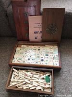 Top view of two nested wooden boxes with Mahjong tiles and sticks visible, plus a pink instruction booklet between them. The tiles show various colorful Chinese symbols.
