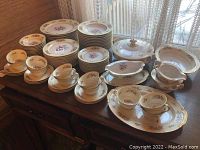 Full set of vintage Noritake china including plates, cups, saucers, a covered soup tureen, creamer, and serving platters arranged on wooden table, showing floral and gold trim design.