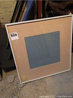 Single large square metal picture frame with white outer mat and central cutout for photo, resting against a wall on the floor