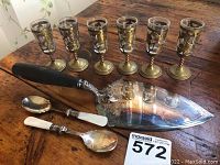Photo showing 12-inch ornate silver-plated cake server with wooden handle, two mother of pearl sauce spoons, and five decorative metal cordial cups in gold-tone holders.