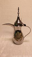 Front angled view of the silver-tone dallah coffee pot showing handle, spout, and floral decoration on the body.
