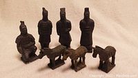 Photo showing all 4 Asian terracotta warrior figurines and 3 horse replicas in one group on a beige fabric background.