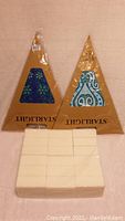 Photo shows two unopened Starlight triangular candles with decorative blue and green pattern inserts standing behind six rectangular bricks of white candle wax wrapped in plastic.