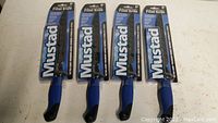 Four unopened Mustad 8-inch fillet knives in original retail packaging positioned horizontally, showing the blue/black handles and blade packaging.
