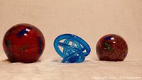 Front view of two red glass spherical paperweights and blue glass knot sculpture against neutral background