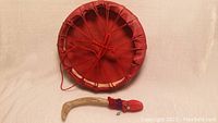 Front view of large red dyed cowhide hand drum with attached red lacing on back, and wooden drumstick decorated with red cloth and beads.