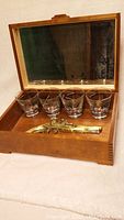 Open hardwood box with mirror lid containing four shooter glasses and a brass pistol wall art