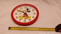 Front view of the 1970s Lorus Mickey Mouse clock showing clock face and Mickey in center. Measuring tape indicates approx. 10 inch diameter.
