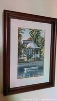 Framed watercolor painting showing a row boat on a waterway in front of a building with greenery, signed at bottom right