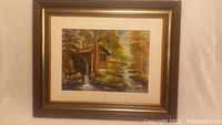 Photo of framed original watercolor painting of a rustic cabin with water wheel next to a flowing stream in a forest with fall foliage.