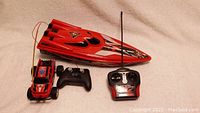 Photo of red remote control boat and red remote control truck, each with their controller, placed on carpeted floor.