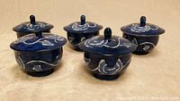 Five small blue ceramic rice bowls with matching lids, featuring white floral and leaf designs. Shown arranged on a beige surface.