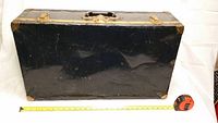 Front view of black steel case trunk showing normal wear scratches, with measuring tape for size reference.