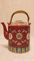 Front view of a vintage Chinese porcelain red Mun Shou longevity teapot showing spout, intricate white floral and Mun Shou emblem pattern on red background, bamboo handle and colorful geometric base.