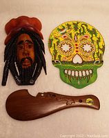 Three wooden art items on white background including carved wooden Patu paddle, 3D carved Rasta head, and hand painted sugar skull.