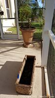 Wide photo showing the urn-shaped plastic planter containing a live palm tree and the adjacent rectangular concrete planter on a patio floor.