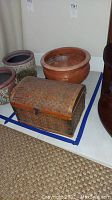 Photo showing large clay pot with reddish-brown color and small woven trunk with latch placed on floor.