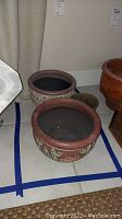 Three clay plant pots on a tiled floor. A brown pot with raised green and white leaf design is in front, beige pot with decorative textures and maroon colors behind, small plain green pot also visible.