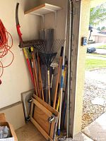 Cluster of garden tools such as rakes, brooms, shovel, hoe, various handles, and a dolly leaning against a garage wall.