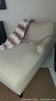 Side angled view of beige lounge chair with throw blanket laid over it, showing chaise design and fabric texture.