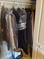 Photo of multiple men's jackets and blazers hung on a clothing rod, some covered in plastic garment bags. Shows variety of colors and materials.