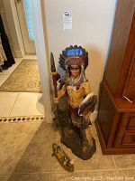 Full view of a standing Native American warrior figurine wearing a feather headdress, holding a spear and shield, placed on the floor next to a piece of furniture.