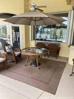 Wide shot showing patio umbrella open over a glass-top round table and wicker chairs on a large brown outdoor rug.