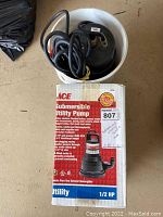 Submersible utility pump inside white bucket with electrical cord and original ACE brand product box next to it