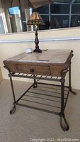 Photo of a square wicker side table with metal legs, a drawer and a tiled top, holding a lamp and other items on top