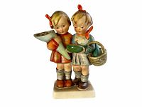 Front view of Hummel figurine showing two children, one holding a cone and the other holding a green bowl with basket on back, on white rectangular base.