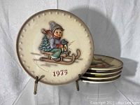Five cream-colored Hummel collectible plates stacked with one plate displayed upright, showing a child figure riding a sled holding a lantern, labeled 1975 in red.