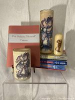 Lot showing two decorative Hummel style candle holders with illustrations of children, a small figurine candle, and a stack of Hummel books in the background.