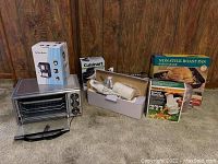 Photo showing the complete lot including Hamilton Beach toaster oven, coffee maker in box, Cuisinart waffle maker, Presto salad shooter, non-stick roast pan, and other appliances boxed or unboxed.