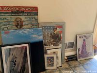 Wide shot revealing ten art pieces stacked against a wall. Includes framed prints and canvases, notably a vintage style satirical poster, a 'New Mexico' print, and others.