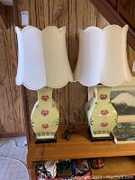 Pair of Asian style ginger lamps sitting on a wooden shelf, showing overall lamps with white scalloped shades and ceramic floral bases.