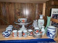 Shelf display of assorted Japanese porcelain items: Imari-style mugs, saucers, serving stands, plates, bowls; blue and white lidded jars and brush pot; white bud vases; floral and green vases