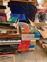 Stacks of hardcover and softcover books in various genres displayed on a concrete floor alongside a fan and boxed kitchen appliance