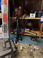 Photo shows tripods, brass telescope-style lens, clocks, watches and camera bags on floor and shelf