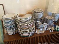 Stack of multiple decorative plates, several mugs stacked, and wooden salad tongs on top. Plates include various sizes with floral Imari Blossoms patterns.