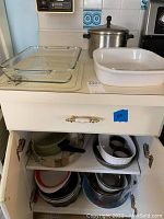 Overview of kitchen countertop showing glass and ceramic lasagna dishes and aluminum stockpot with lid. Cabinet below reveals various frying pans, mixing, and serving bowls stored inside.