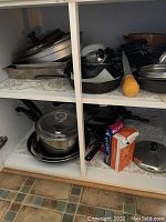 Cabinet shelf displaying stacked aluminum baking dishes, cast iron frying pans, sauce pans, and a yellow-handled juicer.