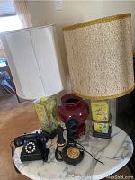 Two yellow ceramic table lamps with floral design, two vintage telephones in front, and red vase behind on a marble-topped table.